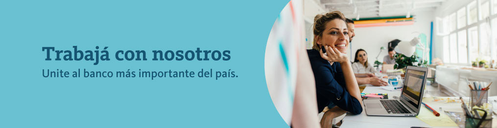 Unite al banco más importante del país imagen de una mujer sonriendo junto a su computadora y sus compañeros de trabajo.